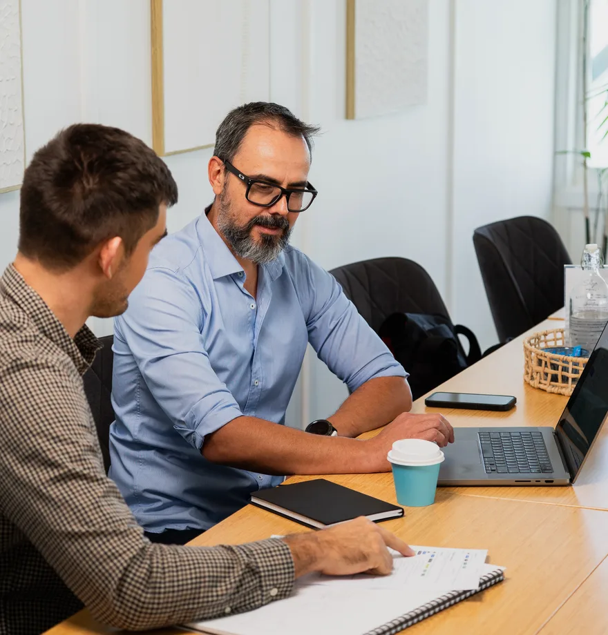 Two engineers working through a problem together at a desk, one explaining while the other takes notes